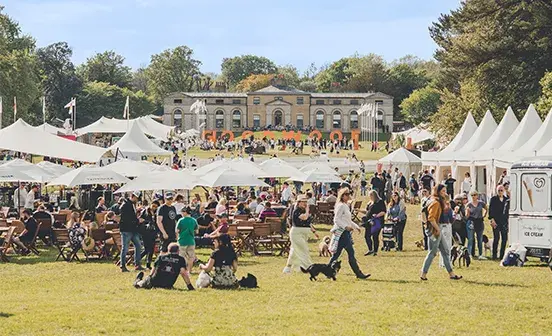 A large outdoor festival scene on a sunny day, with crowds of people sitting at wooden tables and walking across a grassy field. White tents and umbrellas line both sides, with food and vendor stalls visible. In the background, a stately historic building sits on a hill, and large orange letters spell ‘GOODWOOF.’ Many attendees are with dogs, creating a lively, family- and pet-friendly atmosphere.